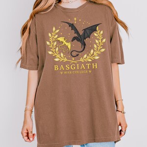 May include: Brown t-shirt featuring a dragon graphic. The design includes a black dragon, a yellow dragon, and the text "BASGIATH WAR COLLEGE" in gold. The dragons are surrounded by a gold wreath.