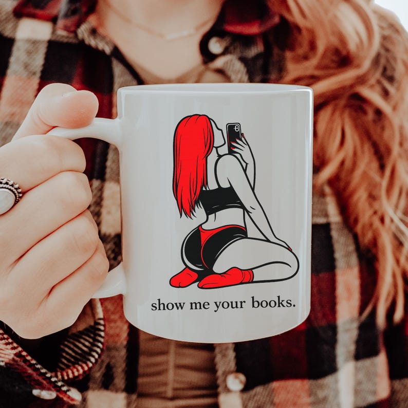 Pu&ograve; includere: Tazza in ceramica bianca con una grafica di una persona con i capelli rossi che si fa un selfie. La persona indossa un top nero, pantaloncini rossi e calzini rossi. Il testo "show me your books" &egrave; stampato sulla tazza.