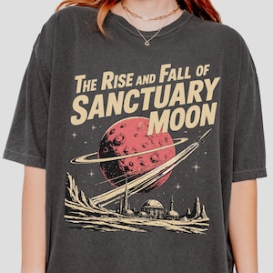 Pode incluir: T-shirt cinza escuro com um gráfico retrô. O design inclui um planeta vermelho com anéis, um foguete e o texto "The Rise and Fall of Sanctuary Moon". O gráfico também retrata uma cidade futurista e uma cordilheira.