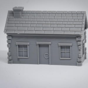 Puede incluir: Una pequeña maqueta gris de una cabaña con chimenea, tejado de tejas y dos ventanas. La cabaña tiene una puerta principal con un pomo redondo. El diseño es una representación en miniatura de una vivienda rústica.