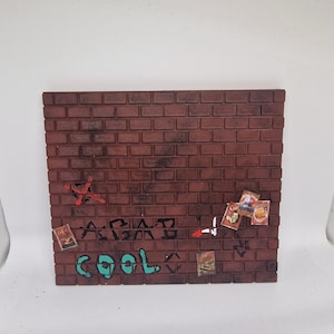 May include: A square, textured red brick wall display piece featuring black graffiti text that reads "AGAB" and "cool" in teal. The surface includes a red painted cross, several small rectangular posters, and weathered dark shading across the bricks.