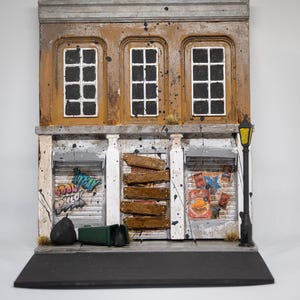 Puede incluir: Diorama en miniatura de una fachada de edificio con tres ventanas arriba y dos escaparates abajo. Las ventanas tienen cristales negros. Un escaparate tiene graffiti que dice "Cool Dillo". El otro tiene un anuncio de hamburguesas. Una farola negra se alza a la derecha.