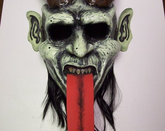 May include: A grotesque mask with a pale green face, black eye sockets, and a long, red tongue. The mask has brown horns, pointed ears, and black hair. The teeth are yellowed, and the overall design is detailed and frightening.