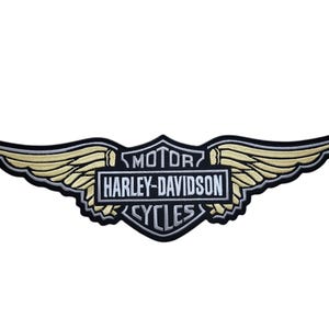 May include: Embroidered patch featuring the Harley-Davidson logo with wings. The design includes the words "MOTOR HARLEY-DAVIDSON CYCLES" in a rectangular frame, with gold and black accents.