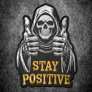 Op de afbeelding: Een zwarte geborduurde patch met een wit skelet in een gewaad met capuchon, dat met beide handen een duim omhoog steekt. Onder het skelet is de tekst STAY POSITIVE in gouddraad geborduurd op de zwarte stoffen achtergrond.