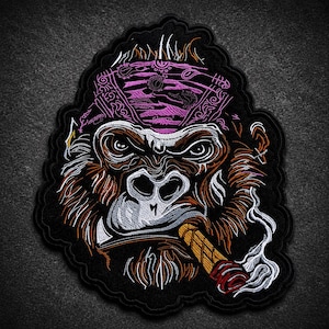 Può includere: Una toppa ricamata raffigurante un gorilla che indossa una bandana rosa a motivi. Il gorilla ha una pelliccia marrone, un muso bianco e fuma un sigaro marrone con fumo bianco che si alza. Il disegno è delineato con filo nero su sfondo nero.
