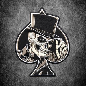 Könnte beinhalten: Ein gestickter Aufnäher in Form eines Pik-Spielkartensymbols mit einem Totenkopf, der einen schwarzen Zylinder und eine Lederjacke trägt. Der Totenkopf ist in Off-White- und Grautönen gehalten, wobei eine Skeletthand nahe der Hutkrempe erhoben ist.