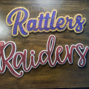 Può includere: Due toppe ricamate con nomi di squadre. La parola "Rattlers" è in blu con un contorno dorato. La parola "Raiders" è in rosso con un contorno bianco. Entrambe le toppe sono su una superficie marrone.