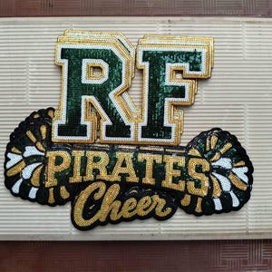 以下が含まれることがあります： ダークグリーンの文字RFにゴールドの縁取りが施されたスパンコールワッペン。その下にゴールドのスパンコールでPIRATES CHEERと記され、両サイドにはグリーン、ゴールド、ホワイトのスパンコールで装飾された2つのポンポンが配置されています。