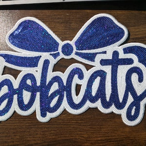 May include: A decorative patch featuring the word Bobcats in a dark blue glitter script font. The text is accented by a large blue glitter bow positioned above the lettering. The entire design is outlined with a thick white border.