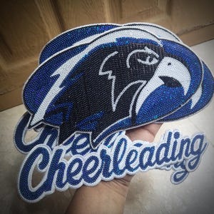 Op de afbeelding: Een set geborduurde patches met een gestileerde adelaarskop van zwarte, witte en blauwe lovertjes, vergezeld van een patch met het woord Cheerleading in blauw cursief schrift met een witte rand en een vulling van blauwe lovertjes.