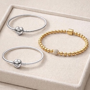 Könnte beinhalten: Drei Armbänder: zwei silberfarbene Armreifen mit runden Verschlüssen, eines mit herzförmigem Anhänger und das andere mit kugelförmigem Anhänger, beide mit dem Wort PANDORA graviert. Das dritte ist ein goldenes Perlenarmband mit einer zentralen Pavé-Perle.