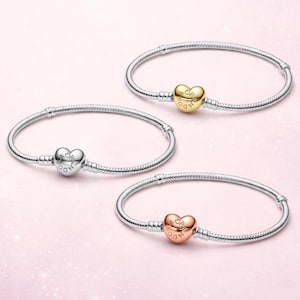 May include: Three snake chain bracelets made of silver-tone metal. Each bracelet features a heart-shaped clasp engraved with the word PANDORA. One clasp is gold-tone, one is silver-tone, and one is rose gold-tone, all arranged on a light pink background.