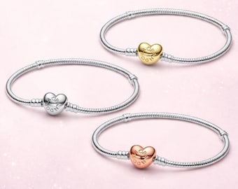 Puede incluir: Tres pulseras de cadena de serpiente de metal plateado. Cada pulsera presenta un cierre en forma de corazón grabado con la palabra PANDORA. Un cierre es dorado, otro plateado y otro oro rosa, dispuestos sobre un fondo rosa claro.