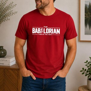 May include: A red short-sleeved t-shirt featuring white text that reads THE BABALORIAN THIS IS THE WAY with a small graphic of a helmet integrated into the letter A. The shirt has a crew neckline and a standard fit, made of solid red fabric.