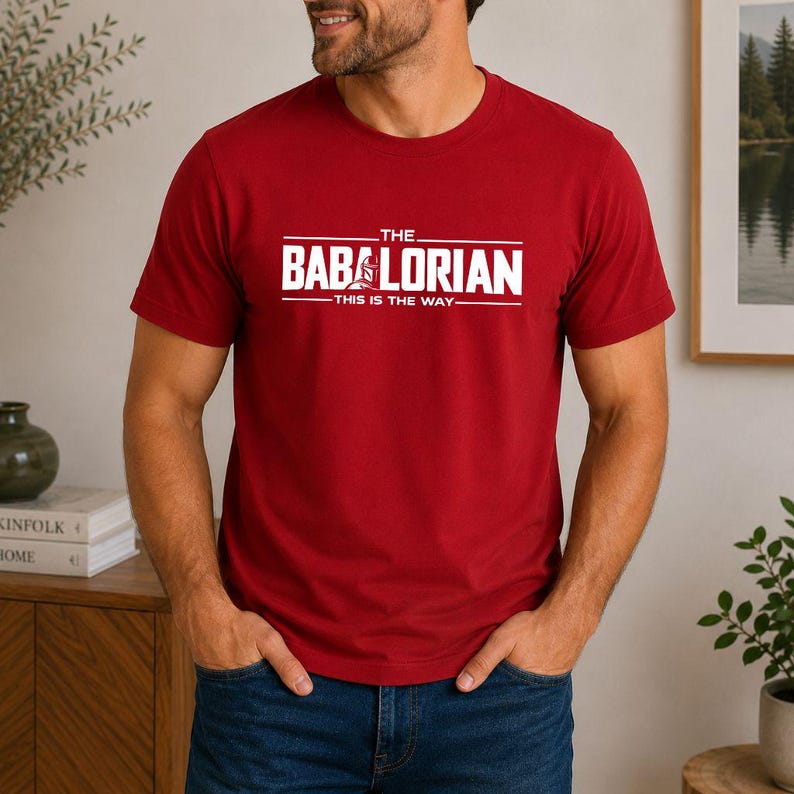 May include: A red short-sleeved t-shirt featuring white text that reads THE BABALORIAN THIS IS THE WAY with a small graphic of a helmet integrated into the letter A. The shirt has a crew neckline and a standard fit, made of solid red fabric.