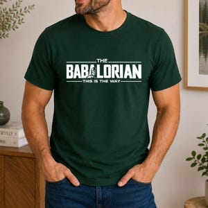 May include: A dark green short-sleeved t-shirt featuring white text that reads THE BABALORIAN THIS IS THE WAY with a graphic of a helmeted character integrated into the lettering. The shirt has a crew neckline and a standard fit design.