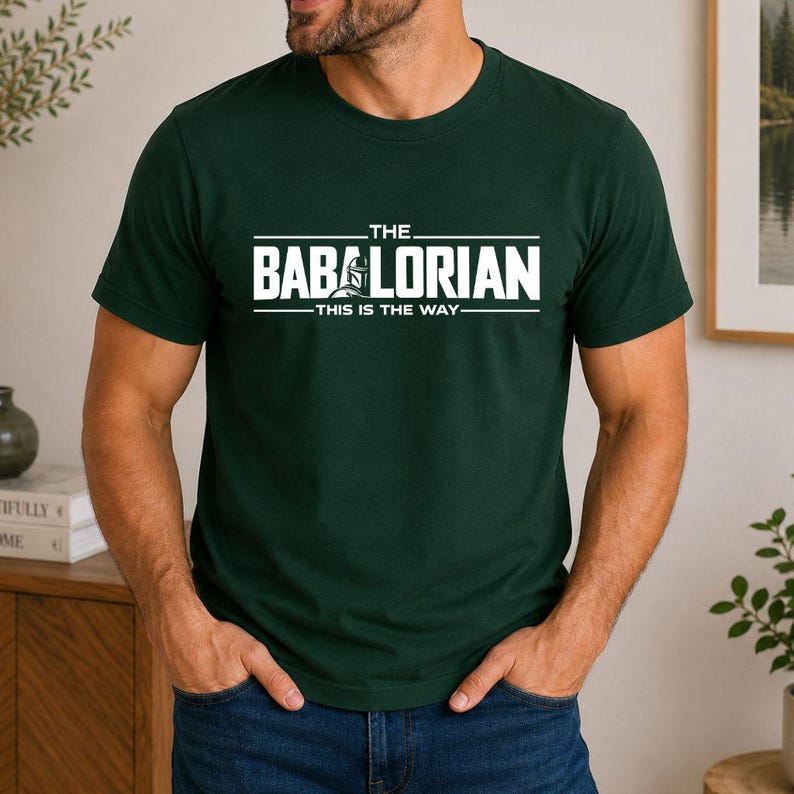 May include: A dark green short-sleeved t-shirt featuring white text that reads THE BABALORIAN THIS IS THE WAY with a graphic of a helmeted character integrated into the lettering. The shirt has a crew neckline and a standard fit design.