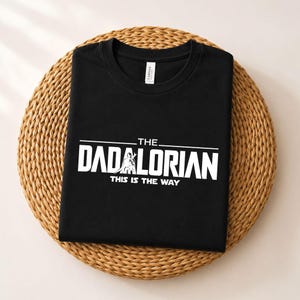 May include: A black short-sleeved t-shirt featuring white screen-printed text that reads THE DADALORIAN THIS IS THE WAY, with a graphic of a figure integrated into the lettering. The shirt has a crew neckline and a visible brand label at the collar.