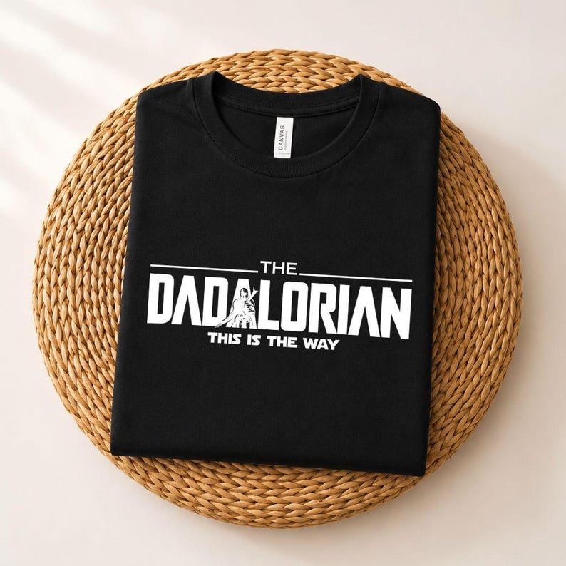 May include: A black short-sleeved t-shirt featuring white screen-printed text that reads THE DADALORIAN THIS IS THE WAY, with a graphic of a figure integrated into the lettering. The shirt has a crew neckline and a visible brand label at the collar.