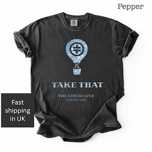 May include: A charcoal gray short-sleeve t-shirt featuring a light blue glittery hot air balloon graphic with a central symbol. Below the balloon, the text reads TAKE THAT, THE CIRCUS LIVE, and SUMMER 2025 in a light blue font on the chest.