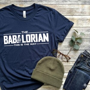 May include: A navy blue short-sleeve t-shirt featuring white screen-printed text that reads THE BABALORIAN THIS IS THE WAY, with a graphic of a helmet replacing the letter A. The shirt is displayed alongside a green beanie and brown glasses.