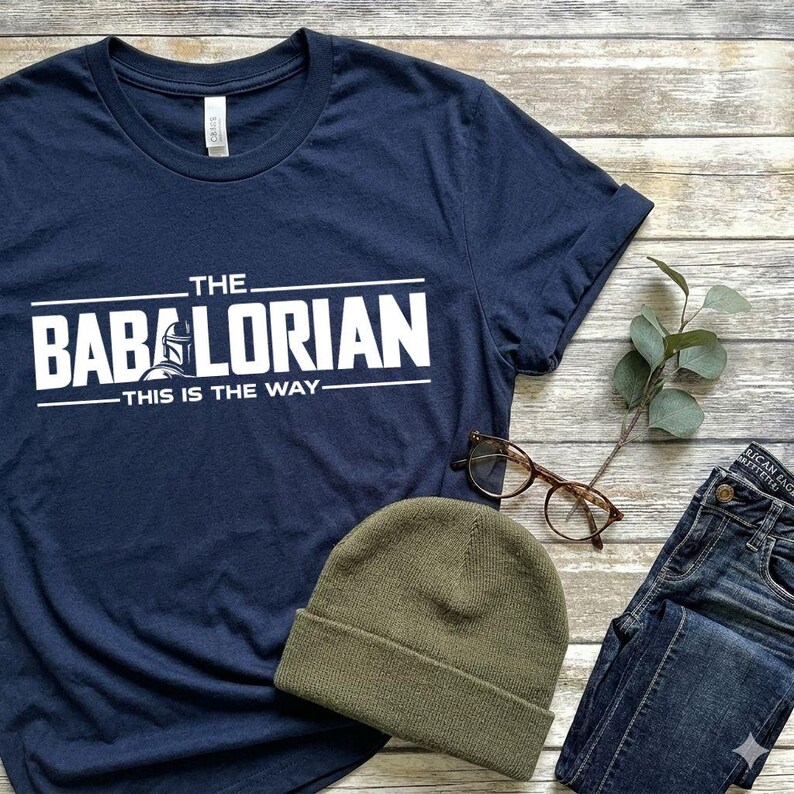 May include: A navy blue short-sleeve t-shirt featuring white screen-printed text that reads THE BABALORIAN THIS IS THE WAY, with a graphic of a helmet replacing the letter A. The shirt is displayed alongside a green beanie and brown glasses.