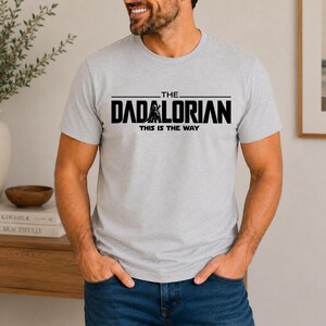 May include: A light gray short-sleeved t-shirt featuring black text that reads THE DADALORIAN THIS IS THE WAY, with a graphic of a helmeted figure integrated into the lettering. The shirt has a crew neckline and a standard fit.