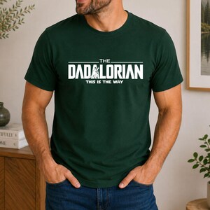 May include: A dark green short-sleeved t-shirt featuring white text that reads THE DADALORIAN THIS IS THE WAY, with a graphic of a figure standing in the center of the word DADALORIAN. The shirt has a crew neckline and a standard fit.