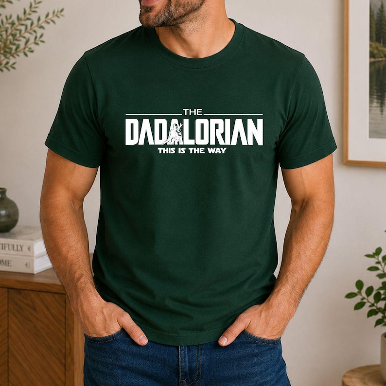 May include: A dark green short-sleeved t-shirt featuring white text that reads THE DADALORIAN THIS IS THE WAY, with a graphic of a figure standing in the center of the word DADALORIAN. The shirt has a crew neckline and a standard fit.