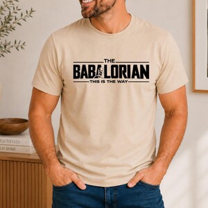 May include: A beige short-sleeved crew neck t-shirt featuring black text that reads THE BABALORIAN THIS IS THE WAY with a small graphic of a helmeted figure integrated into the lettering on the chest area of the garment.