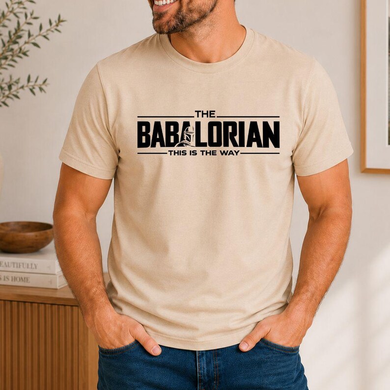 May include: A beige short-sleeved crew neck t-shirt featuring black text that reads THE BABALORIAN THIS IS THE WAY with a small graphic of a helmeted figure integrated into the lettering on the chest area of the garment.