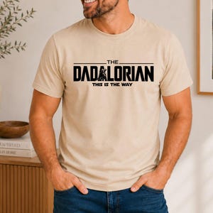 May include: A beige short-sleeved t-shirt featuring black text that reads THE DADALORIAN and THIS IS THE WAY, with a small black graphic of a helmeted figure integrated into the lettering on the chest of the garment.