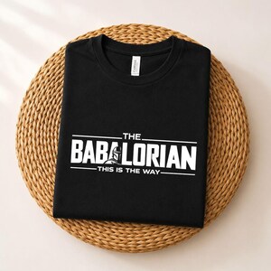 May include: A black crew neck t-shirt featuring white screen-printed text that reads THE BABALORIAN THIS IS THE WAY, with a small graphic of a helmeted character integrated into the letter A. The shirt has a white interior neck label.