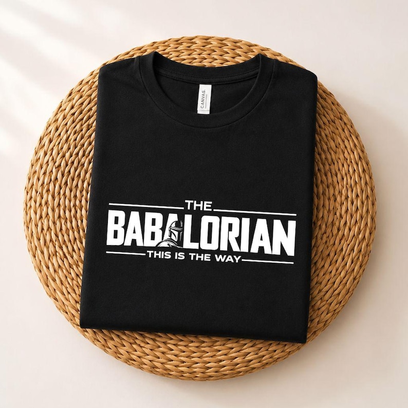 May include: A black crew neck t-shirt featuring white screen-printed text that reads THE BABALORIAN THIS IS THE WAY, with a small graphic of a helmeted character integrated into the letter A. The shirt has a white interior neck label.