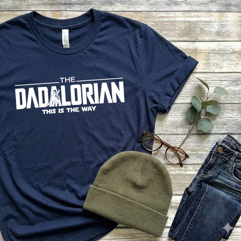May include: A navy blue short-sleeve t-shirt featuring white screen-printed text that reads THE DADALORIAN THIS IS THE WAY, with a small graphic of a figure integrated into the letter A. The shirt is displayed alongside a green beanie and glasses.
