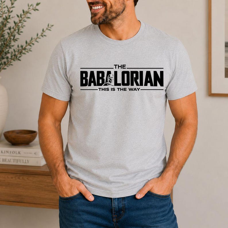 May include: A light gray short-sleeved t-shirt featuring black text that reads THE BABALORIAN with a graphic of a helmet integrated into the letter A and the text THIS IS THE WAY printed in a smaller font directly underneath the main title.