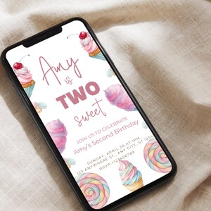 Puede incluir: Un teléfono inteligente muestra una invitación de cumpleaños con ilustraciones de acuarela de cupcakes, algodón de azúcar y una piruleta. El texto dice "Amy is TWO Sweet" con detalles del evento. El teléfono está sobre una tela beige.