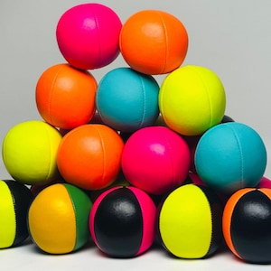May include: A pyramid of colorful juggling balls in shades of pink, orange, teal, and yellow. Some balls are two-toned with black and a bright color. The balls are made of a soft material and are designed for juggling.