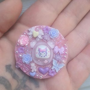 May include: A round, pink, decorative pin with a cartoon character in the center. The pin is adorned with small flowers, hearts, bows, and beads in shades of pink, purple, and white. The pin is held in a hand.