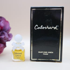May include: A miniature perfume bottle of Cabochard by Grès Paris, with a white bow and a clear bottle filled with yellow liquid. The bottle is next to a black box with gold trim and the Cabochard logo. The bottle contains 1.8 ml.