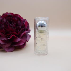 May include: A small perfume bottle with a silver cap is encased in a clear, cylindrical container. A deep burgundy flower is to the left of the bottle. The background is a solid, light beige color.