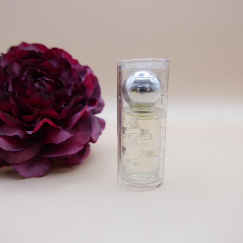 May include: A small perfume bottle with a silver cap is encased in a clear, cylindrical container. A deep burgundy flower is to the left of the bottle. The background is a solid, light beige color.