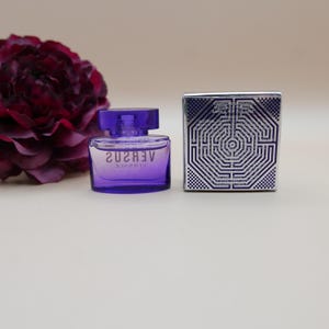 May include: A small purple glass perfume bottle with a matching cap and the text VERSUS VERSACE printed on the front. Beside it sits a square silver box featuring a geometric maze pattern and printed text on the front surface.
