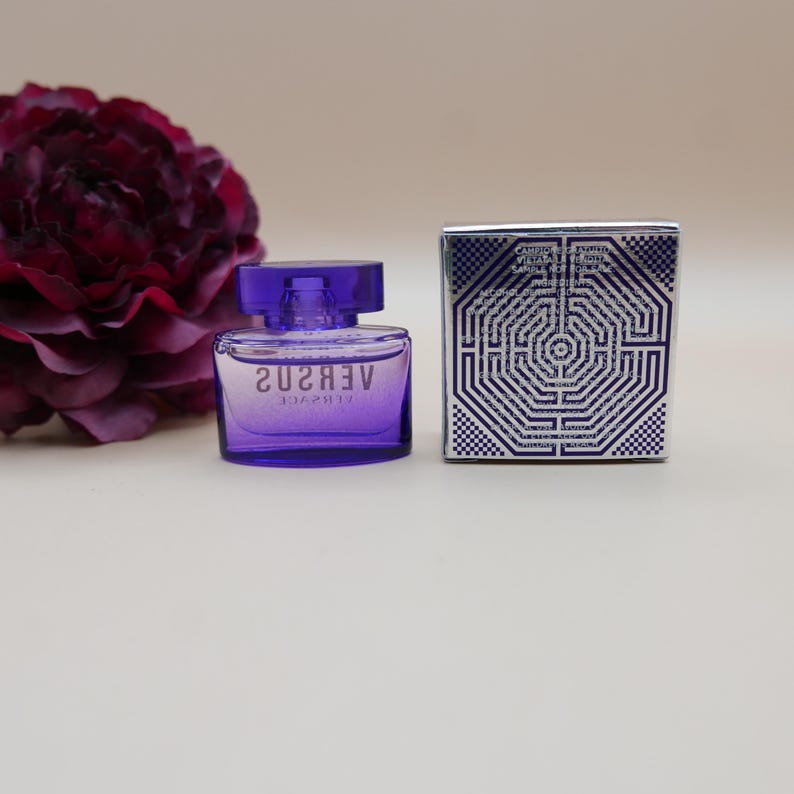 May include: A small purple glass perfume bottle with a matching cap and the text VERSUS VERSACE printed on the front. Beside it sits a square silver box featuring a geometric maze pattern and printed text on the front surface.