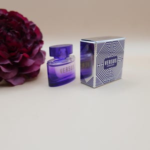 May include: A purple glass perfume bottle with a matching cap and the text VERSUS printed on the front, positioned next to its rectangular box featuring a white and blue geometric maze pattern and the text VERSUS VERSACE on the front panel.