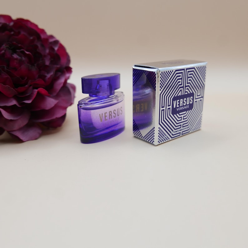 May include: A purple glass perfume bottle with a matching cap and the text VERSUS printed on the front, positioned next to its rectangular box featuring a white and blue geometric maze pattern and the text VERSUS VERSACE on the front panel.