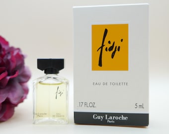 May include: A small glass bottle of Fidji Eau de Toilette with a black cap, positioned next to its white rectangular box. The box features a yellow square with the black text Fidji, the label .17 FL OZ. 5 mL, and the brand name Guy Laroche Paris.