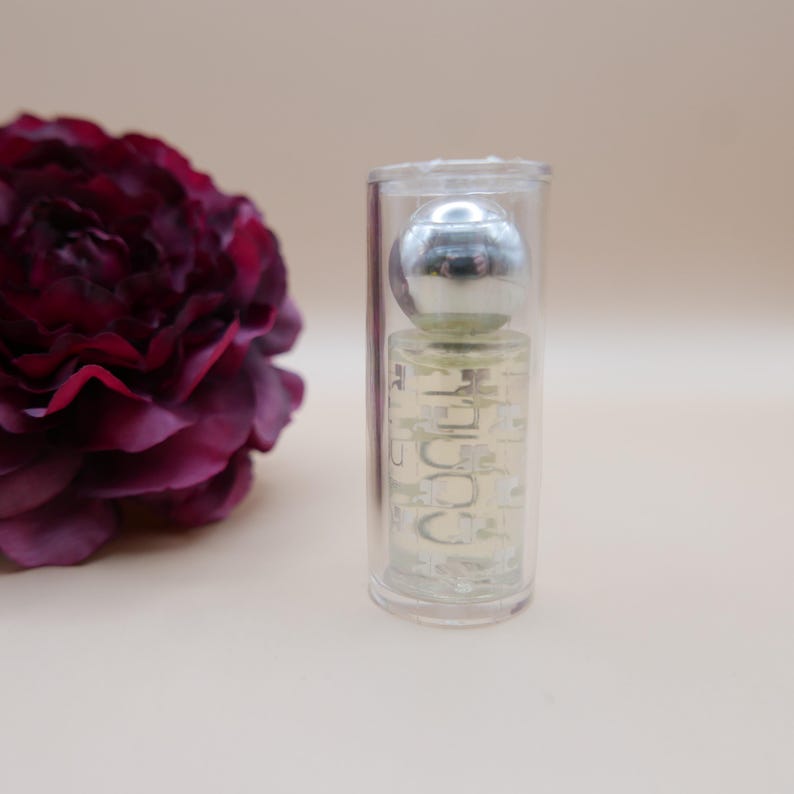 May include: A clear perfume bottle with a silver spherical cap is encased in a transparent tube. The bottle contains a light-colored liquid and has the word "COCO" printed on it. A dark red flower is in the background.