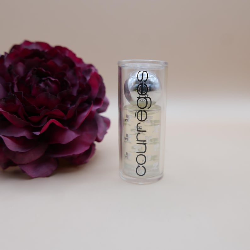 May include: A clear cylindrical perfume bottle with a silver ball top and the word "courr&egrave;ges" printed on it. A dark burgundy flower is to the left of the bottle. The background is a light beige color.
